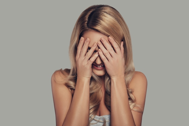 Time to hide. Beautiful young woman covering face with hands and smiling while standing against grey background