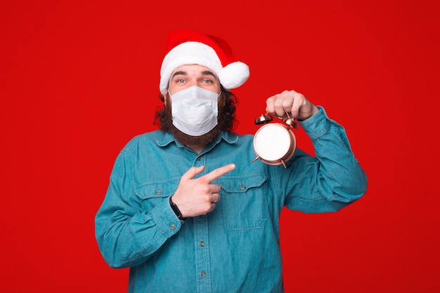 Time for holidays and protection, bearded man with Santa hat and protection mask pointing at clock