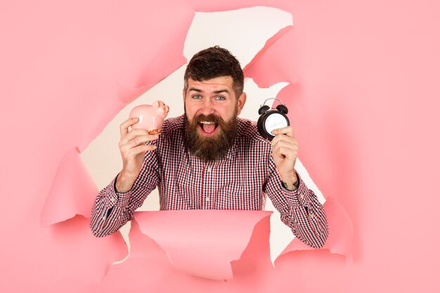 Time is money concept happy bearded man looking through hole in pink paper holds piggybank and clock