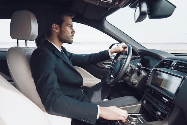 Time is money. Handsome young man in full suit looking straight while driving a car