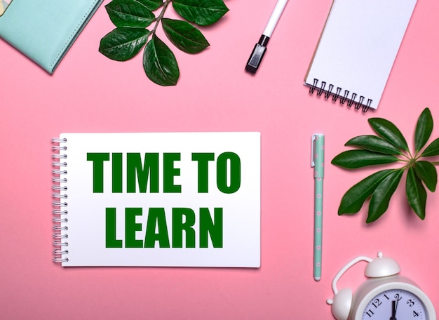 TIME TO LEARN is written in green on a white notepad on a pink wall surrounded by notepads, pens, white alarm clock and green leaves. Educational concept