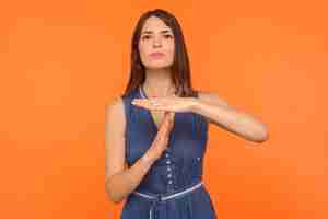 Photo time limit. unhappy brunette woman in denim dress looking with serious beseeching expression and showing time out hand gesture, upset about deadline. indoor studio shot isolated on orange background