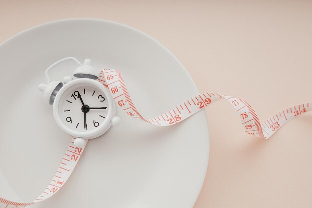 Time to lose weight concept white alarm clock on a white plate\
and measuring tape closeup