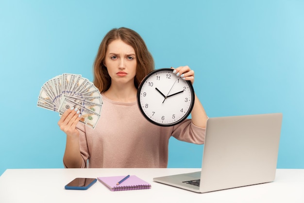 Time to make money. Strict bossy confident woman sitting at workplace, holding clock and dollar bills, looking at camera with angry defiant expression. indoor studio shot isolated on blue background