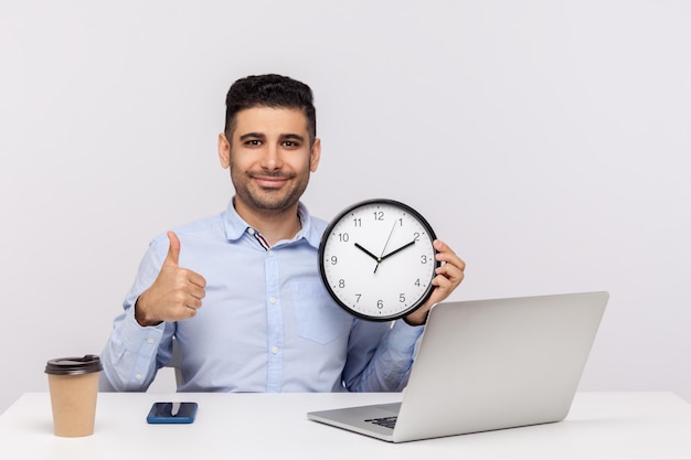 Тайм-менеджмент. Веселый молодой человек сидит на рабочем месте с ноутбуком, держит большие часы и показывает большой палец вверх, как жест, улыбаясь в камеру. студийный снимок на белом фоне