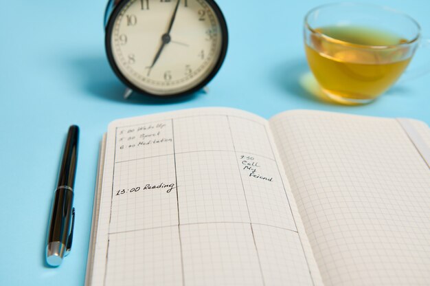 Time management, deadline and schedule concept: alarm clock on schedule plan, organizer with plans, ink pen and transparent glass cup with tea on blue background with space for text