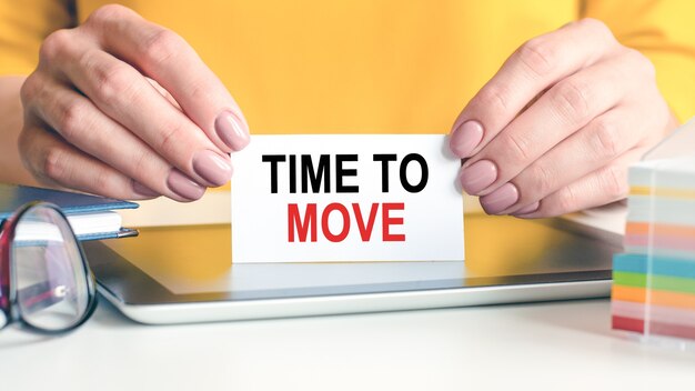 Time to move is written on a white business card in a woman's hands. Yellow background. Glasses, tablet and block with multi-colored paper for notes. Can be used for business, advertising concept.