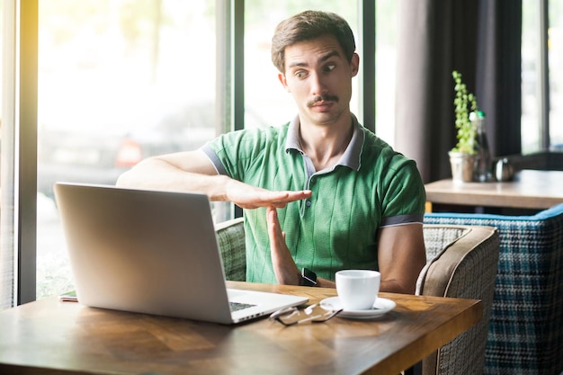 Тайм-аут. Молодой бизнесмен в зеленой футболке работает, смотрит на экран ноутбука во время видеозвонка и умоляет потратить больше времени. концепция бизнеса и фриланса. выстрел в помещении возле окна в дневное время.