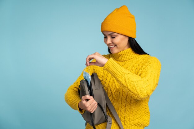 Time for protective mask. smiling brunette woman wearing yellow\
knitted sweater and hat taking off from her backpack her protective\
mask while posing in the studio. pandemic concept