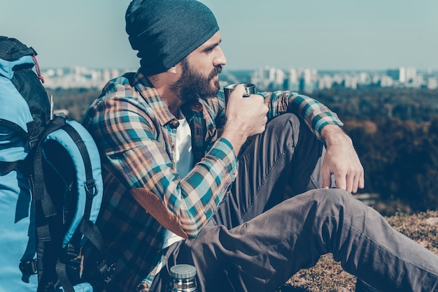 Time to relax. Handsome young man sitting near backpack and looking away while drinking