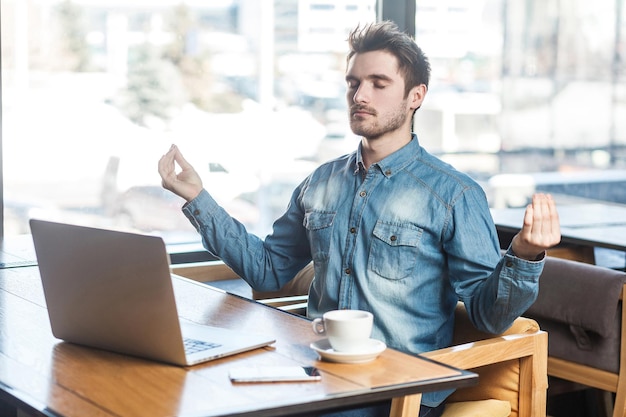 Time to relax! Portrait of handsome successful bearded young freelancer in blue jeans shirt are sitting in cafe and having a rest, holding hands like have meditative to have emotional pleasure.