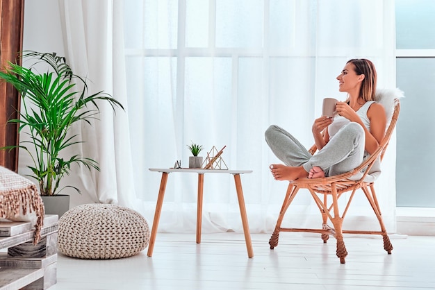 Photo time relax smiling girl drinking hot tea sitting in wicker chair and looking out of window at home