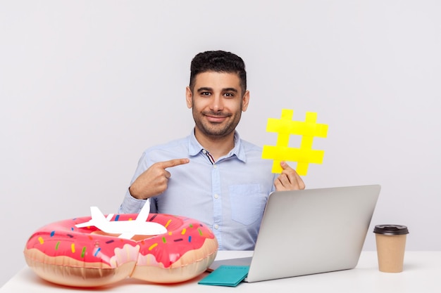 Time to rest. happy man pointing hashtag symbol, sitting in
office workplace with rubber ring passport and paper airplane on
desk. showing hash sign of viral web topic about relax travel.
studio shot