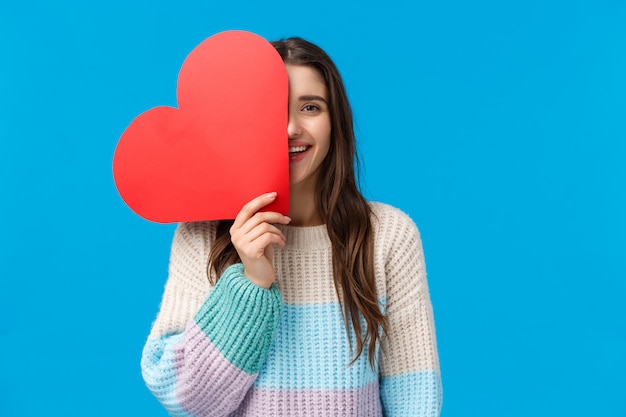 È ora di dire che ti amo. allegro sognante e carino caucasico donna bruna copre metà viso con grande segno di cuore rosso, sorridente, affetto espresso e simpatia il giorno di san valentino, ricevi un regalo carino