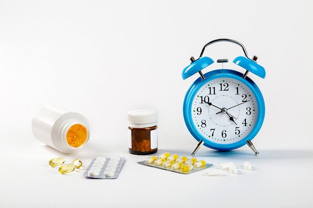 Time to take your pills. An alarm clock on a white wall shows the time of taking medication, and next to it - medical pills.