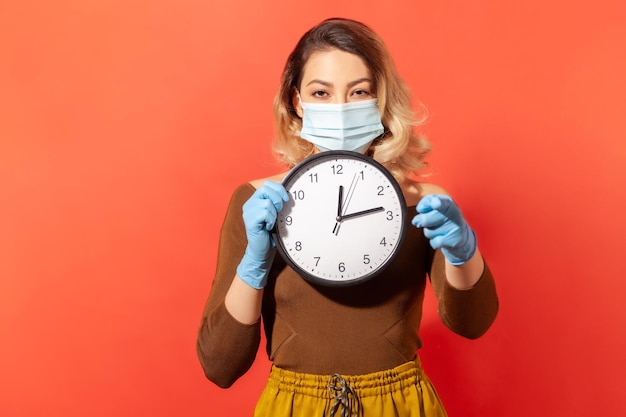 Фото Время оставаться дома женщина в хирургической маске и перчатках, указывающая на камеру и держащая часы, означающая карантинную самоизоляцию, в то время как заразная болезнь распространяется, вспышка коронавируса изолирована в помещении