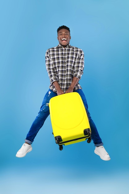 Time to travel full length of excited young black man jumping with bright suitcase over blue studio