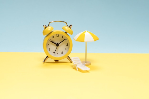 Time to travel. Sun lounger, yellow umbrella and alarm clock on a yellow, blue background.