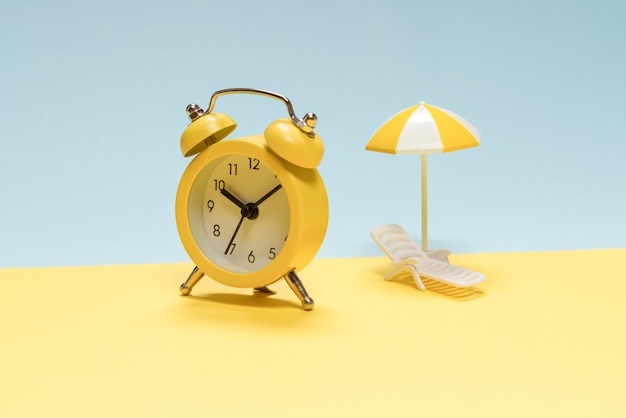 Time to travel. Sun lounger, yellow umbrella and alarm clock on a yellow, blue background.