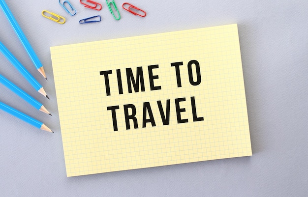 TIME TO TRAVEL text in notebook on gray background next to pencils and paper clips