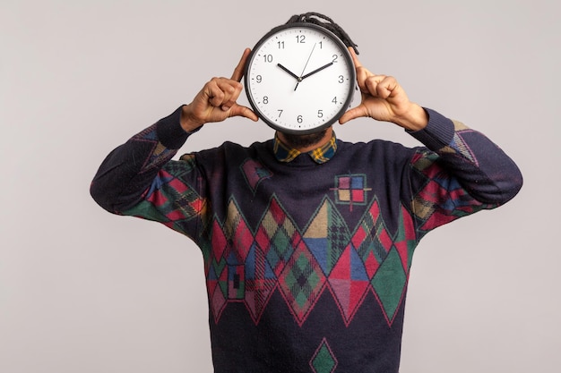 Time to work. African man hiding face behind big wall clock, afraid of deadline, wasting time, being late. Indoor studio shot isolated on gray background