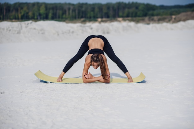 Time for yoga. Attractive and healthy young woman doing exercises on the beach at sunrise or sunset. Fitness girl doing stretching on yoga mat.