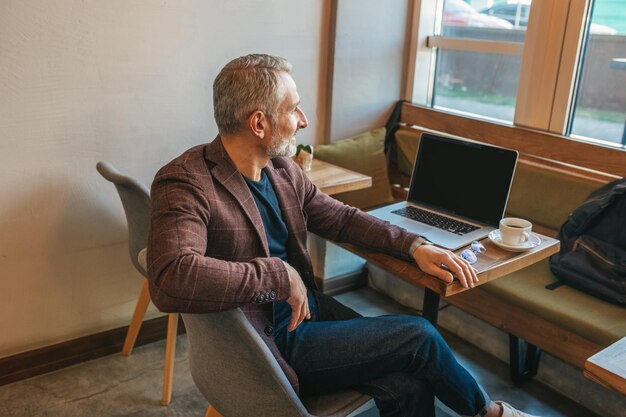 Time for yourself. gray-haired elegant man relaxed sitting at\
table with laptop and coffee thinking looking out window