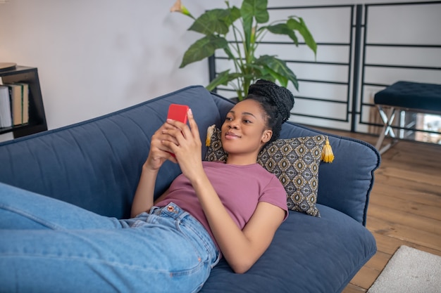 Time for yourself. Pretty young adult slender african american woman with hairstyle looking interested in smartphone lying on sofa