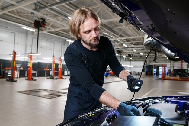 Timely diagnostics of the car at the service A man with a flashlight is looking for malfunctions under the hood of a car