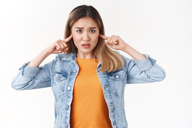 Timid innocent upset asian blond girl cannot stand people fighting shouting frowning nervously look camera shut ears unwilling hear loud noisy party music standing white wall disturbed