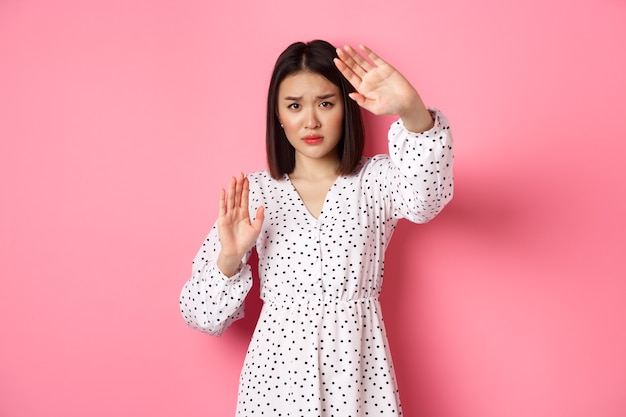 Timid and scared asian woman defending herself, raising arms in protection, victim being attacked, standing over pink background