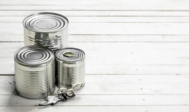 Tin cans with food and opener. On a white wooden table.