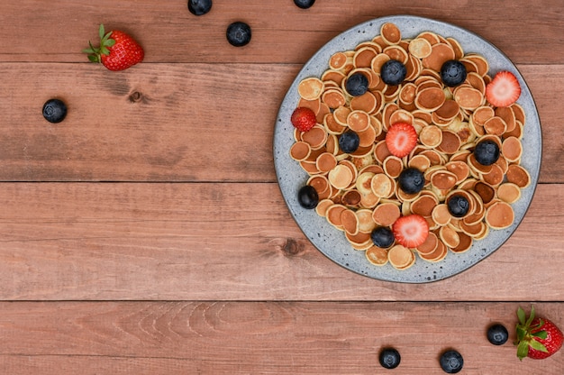Mini pancake dei cereali minuscoli in una ciotola grigia con le fragole ed i mirtilli su fondo di legno.