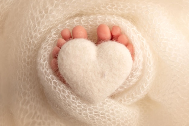 The tiny foot of a newborn baby. Soft feet of a new born in a white wool blanket. Close up of toes, heels and feet of a newborn. Knitted white heart in the legs of a baby. Studio macro photography.