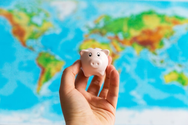 A tiny piggy bank is held in the hand a map of the world in the background