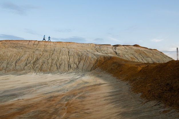 Tiny silhouettes of two contemporary researchers in protective coveralls moving along hill against clear blue sky during investigation