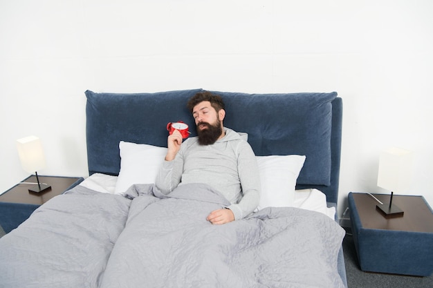 Tips om vroeg wakker te worden Man met baard slaperig gezicht bed met wekker in bed Wat een vreselijk geluid Zet dat rinkelen uit Probleem 's morgens vroeg wakker worden Opstaan met wekker Meer slaap nodig