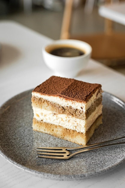 Torta dolce al tiramisù su un piatto con una forchetta e una tazza bianca di caffè nero al tavolo di un caffè messa a fuoco selettiva dell'immagine stile di vita profondità di campo sfondo bokeh