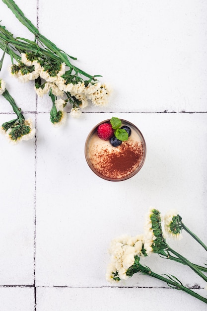 Tiramisu dessert in glass with berries on white tile background with flowers high quality photo