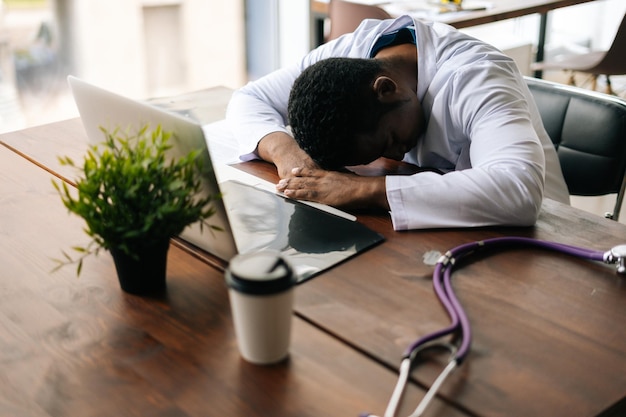 Photo tired african american male doctor fall sleeping at desk on working place. concept of medicine and health care.