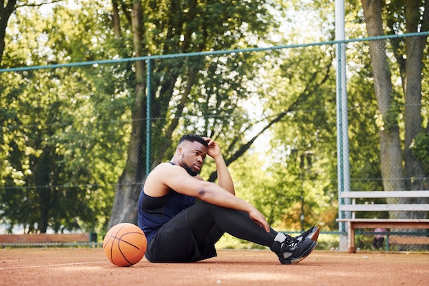 Tired african american man sits on the ground with ball on the court outdoors Takes a break