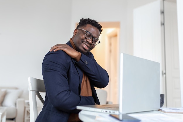 Tired African businessman working from home on his computer feeling pain in his shoulder man suffering from shoulder and back pain while sitting and working from home on laptop