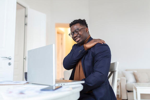 Tired African businessman working from home on his computer feeling pain in his shoulder man suffering from shoulder and back pain while sitting and working from home on laptop