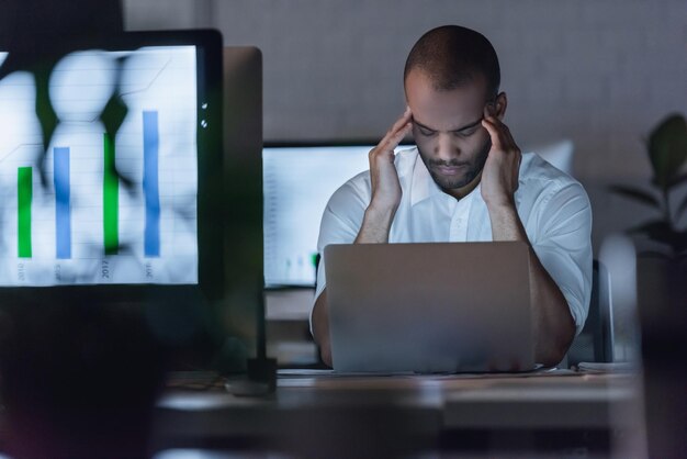 Tired Afro American businessman is massaging his temples while working with a laptop in office late at night