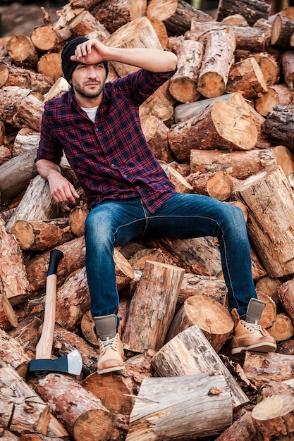 Tired after hard work. Full length of tired young forester touching his forehead while sitting on logs