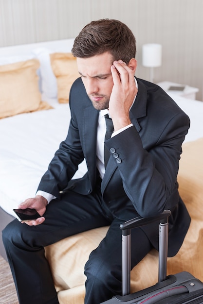 Tired after long business trip. Depressed young businessman in formalwear holding mobile phone and touching head with hand while sitting on the bed in hotel room