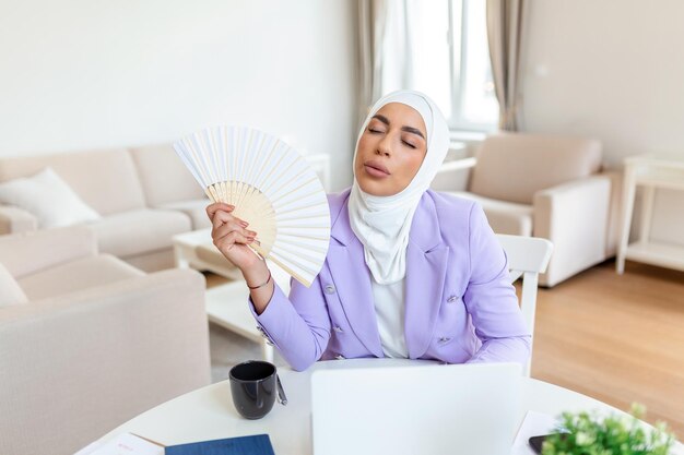 Photo tired arabic muslim woman work on laptop at home office wave with hand fan suffer from heatstroke indoors unwell exhausted young female use waver breathe fresh air lack ventilation conditioner