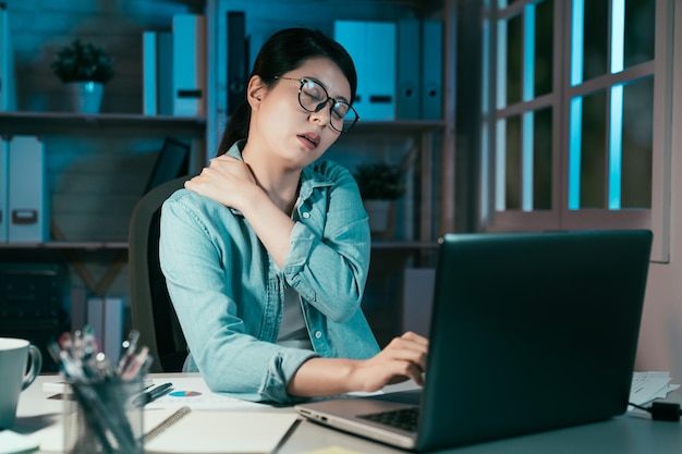 Tired asian chinese businesswoman feeling neck pain working on laptop at night in modern home office. health care concept. young lady worker with painful face and massage shoulder at workplace.