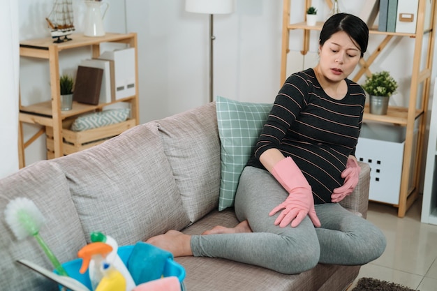 Tired asian expectant pregnant female suffering edema is\
looking at and massaging swollen legs on the sofa while taking a\
rest from housecleaning in living room at home.