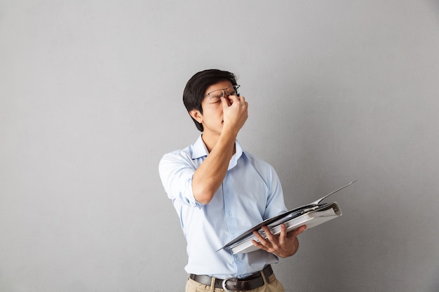 Tired asian man standing isolated, carrying folders with documents